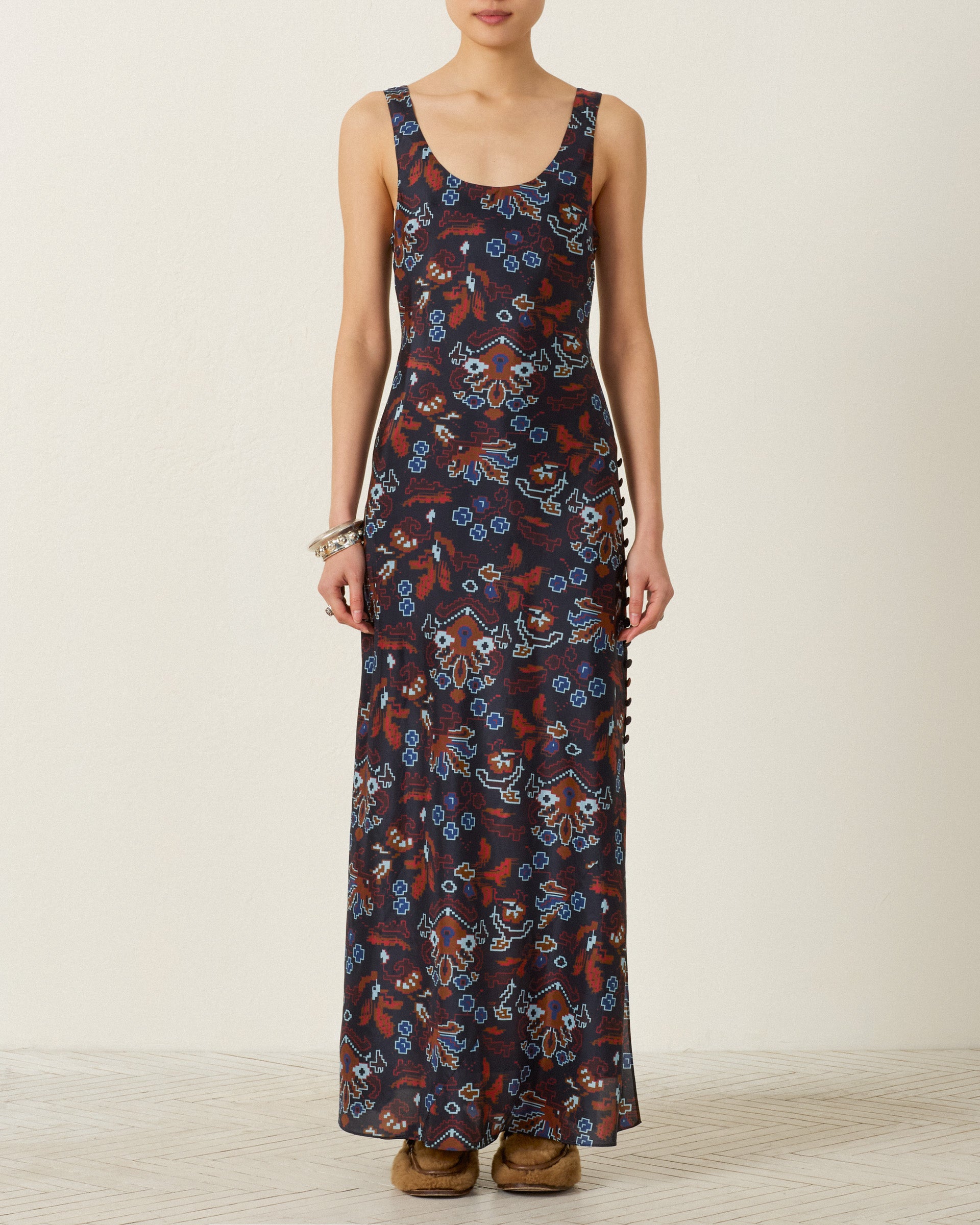 A person wearing the Adele Indigo Kilim Silk Dress—a sleeveless, floor-length hand-loomed silk dress featuring a blue and red floral pattern—stands against a plain off-white background.