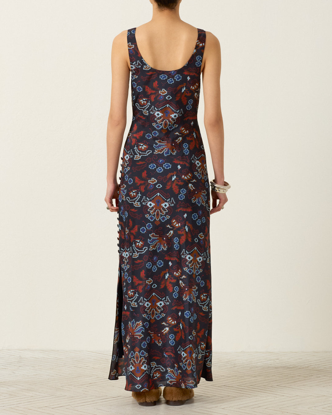 A person stands with their back to the camera, wearing the Adele Indigo Kilim Silk Dress—a sleeveless, floor-length bias-cut slip dress featuring a multicolored floral pattern—paired with chunky bracelets, set against a neutral background.