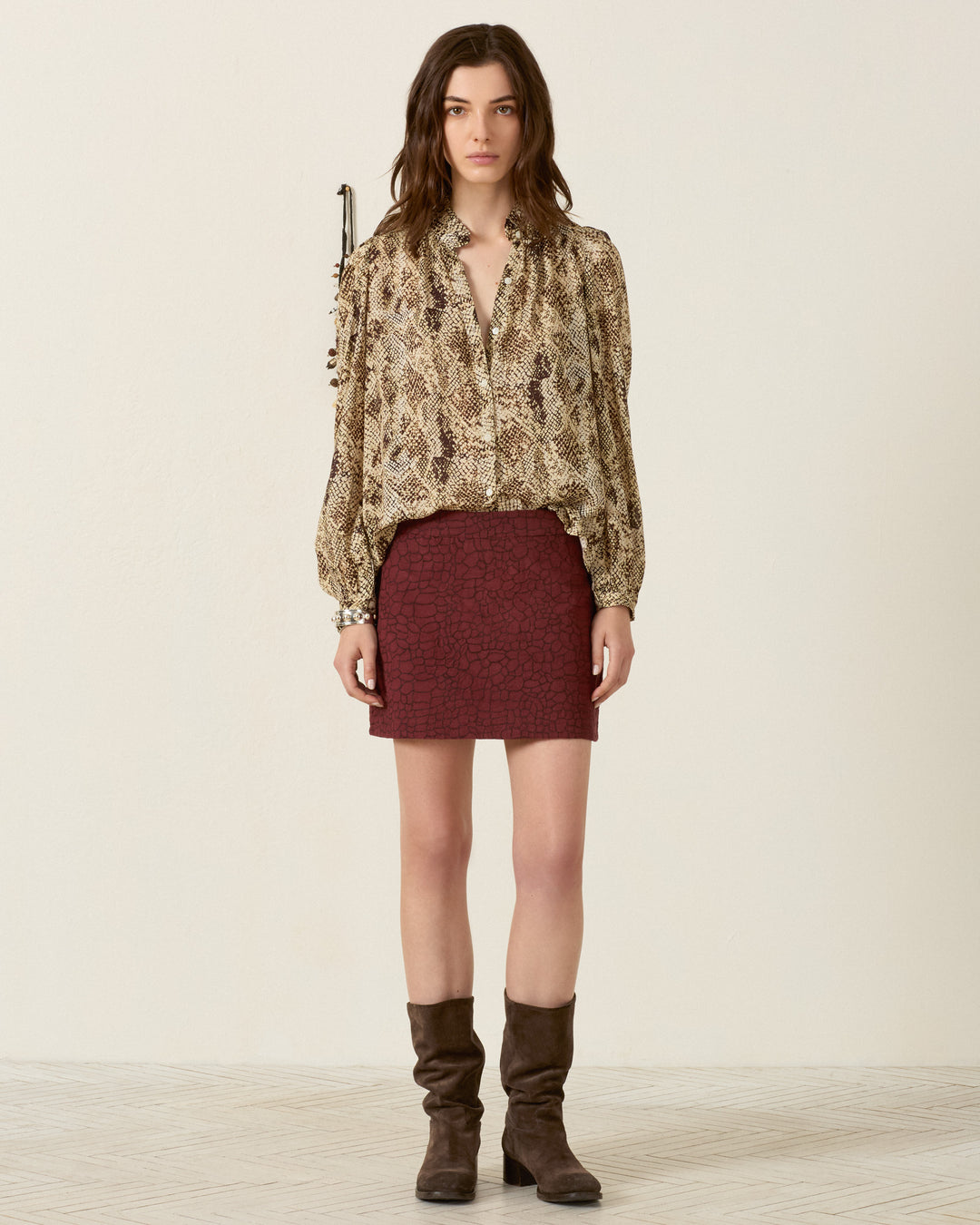A woman stands facing forward, wearing the Annabel Lemon Snakeskin Shirt with puff shoulders, a maroon skirt, and brown knee-high boots, against a plain light-colored background.