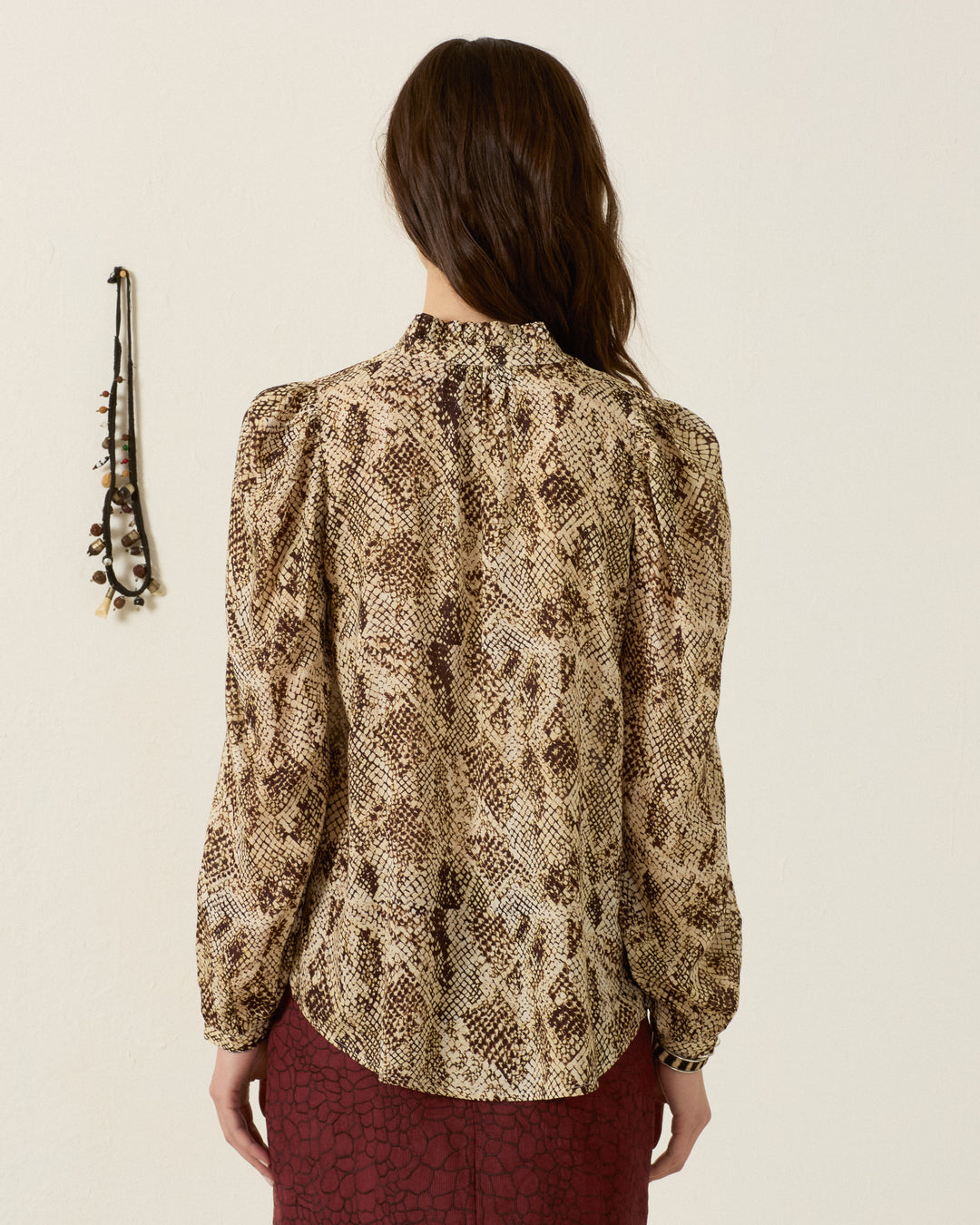 A woman stands facing away, wearing the Annabel Lemon Snakeskin Shirt with long sleeves and puff shoulders, paired with a maroon skirt. A necklace hangs on the wall to her left.