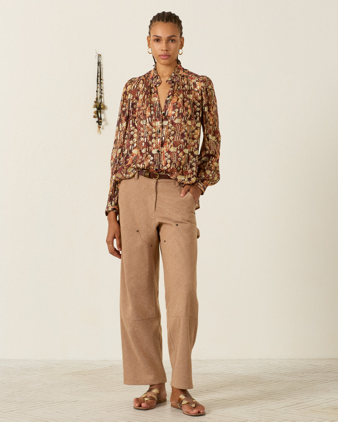A person is standing indoors wearing the Annabel Topaz Snakeskin Shirt with puff shoulders, tan wide-leg pants, and sandals. A decorative wall hanging is visible in the background.