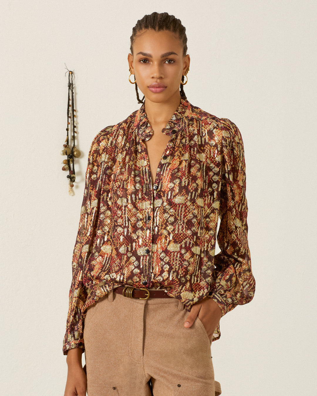 A person with braided hair wears the Annabel Topaz Snakeskin Shirt, featuring puff shoulders, paired with tan pants, standing against a plain wall adorned with hanging beads in the background.
