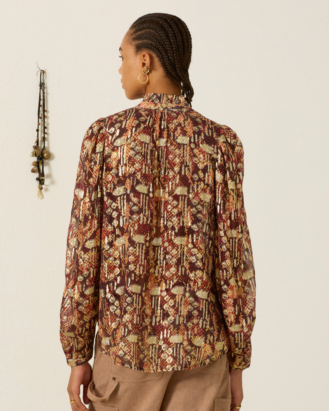 Wearing gold hoop earrings and the Annabel Topaz Snakeskin Shirt—a patterned long-sleeve blouse with a ruffled collar—a person with braided hair faces away from the camera, standing against a plain wall adorned with hanging decor.