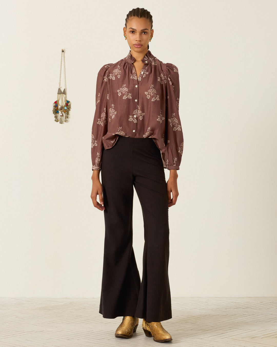 Person standing against a plain wall, wearing the Annabel Doe Elderflower Shirt—a brown floral blouse with a ruffled collar and puff shoulders—paired with black flared pants and gold ankle boots. A keychain holder with keys hangs on the wall to the left.