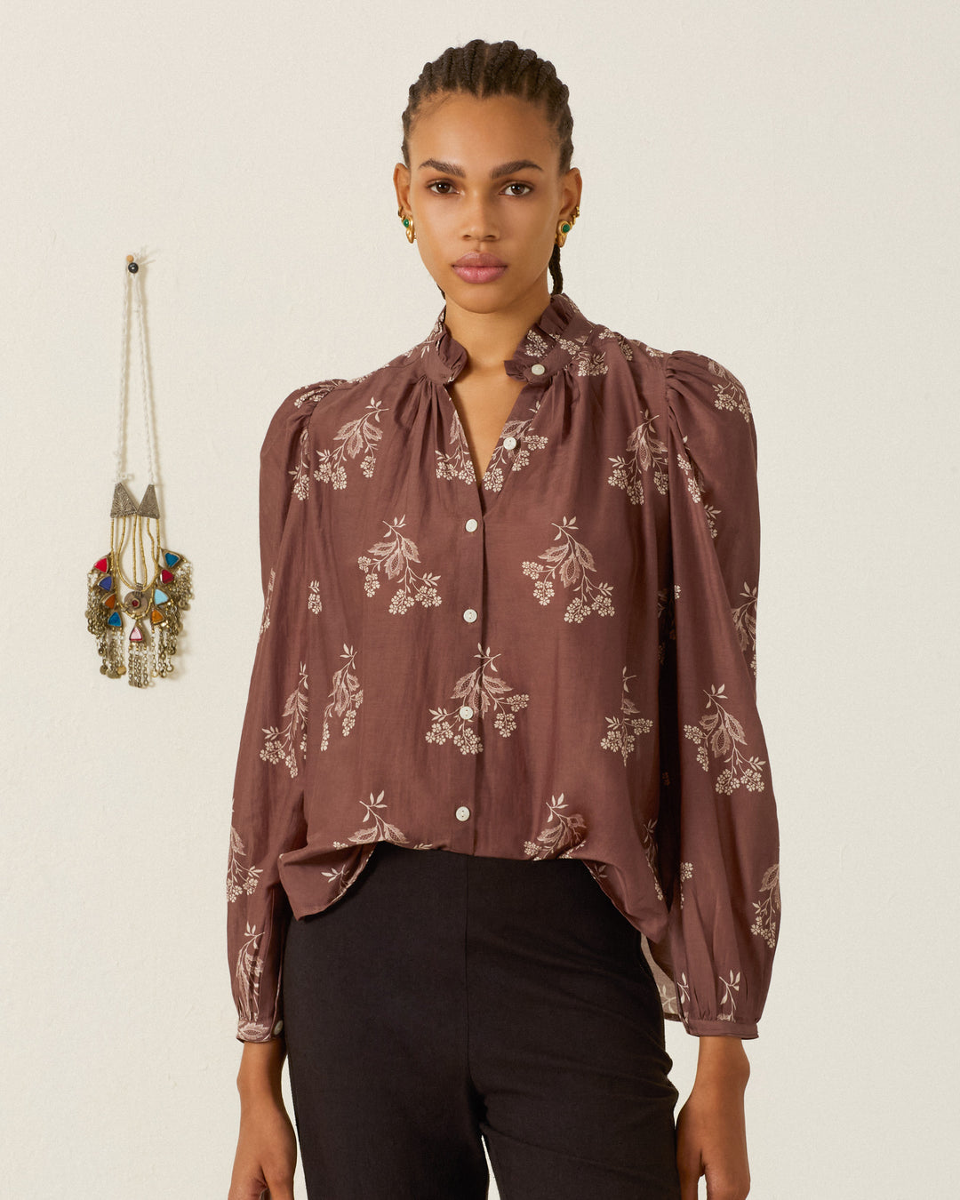 A woman with braided hair wears the Annabel Doe Elderflower Shirt—a brown, long-sleeved floral blouse with puff shoulders—paired with black pants, standing indoors near a wall-mounted decorative item.
