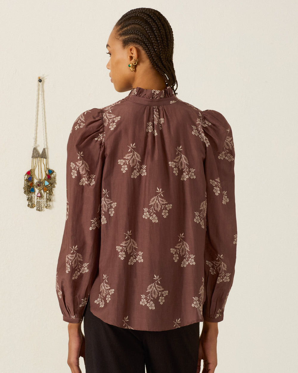 A person with braided hair, wearing the Annabel Doe Elderflower Shirt—a mauve long-sleeve floral blouse with a ruffled collar—and dark pants stands facing away from the camera. A decorative wall hanging is visible on the left.