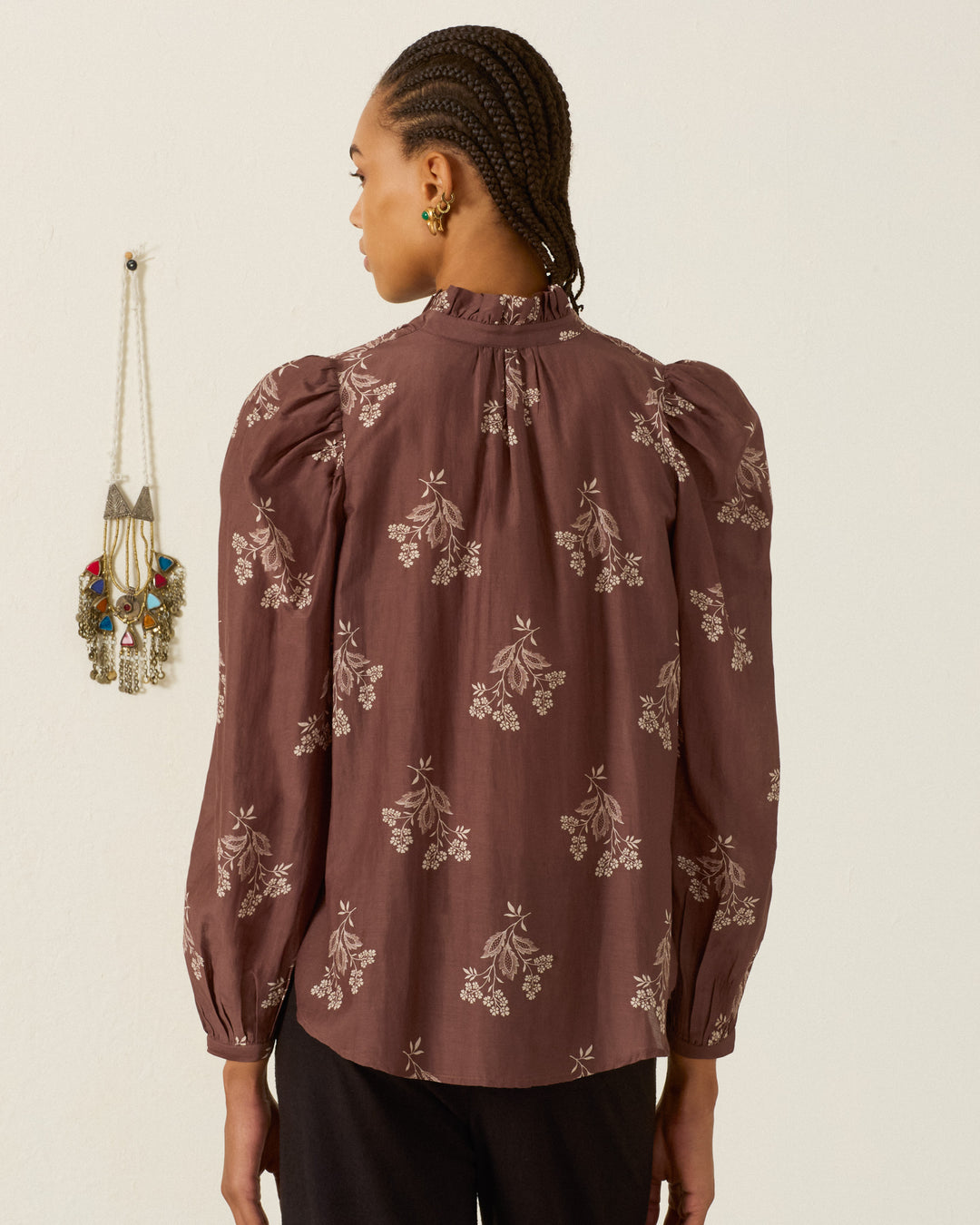 A person with braided hair, wearing the Annabel Doe Elderflower Shirt—a mauve long-sleeve floral blouse with a ruffled collar—and dark pants stands facing away from the camera. A decorative wall hanging is visible on the left.