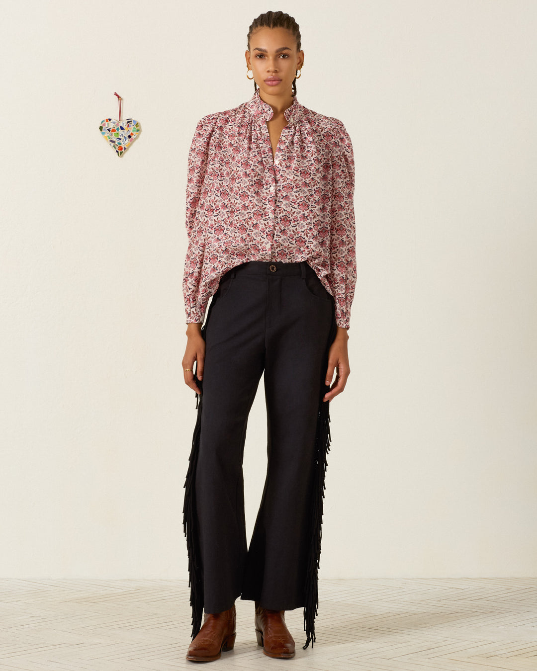 Wearing the Annabel Horizon Kilim Shirt, featuring a vibrant red floral pattern and ruffled collar, along with black fringe pants and brown boots, the person stands against a plain background accented by a small heart ornament on the wall.