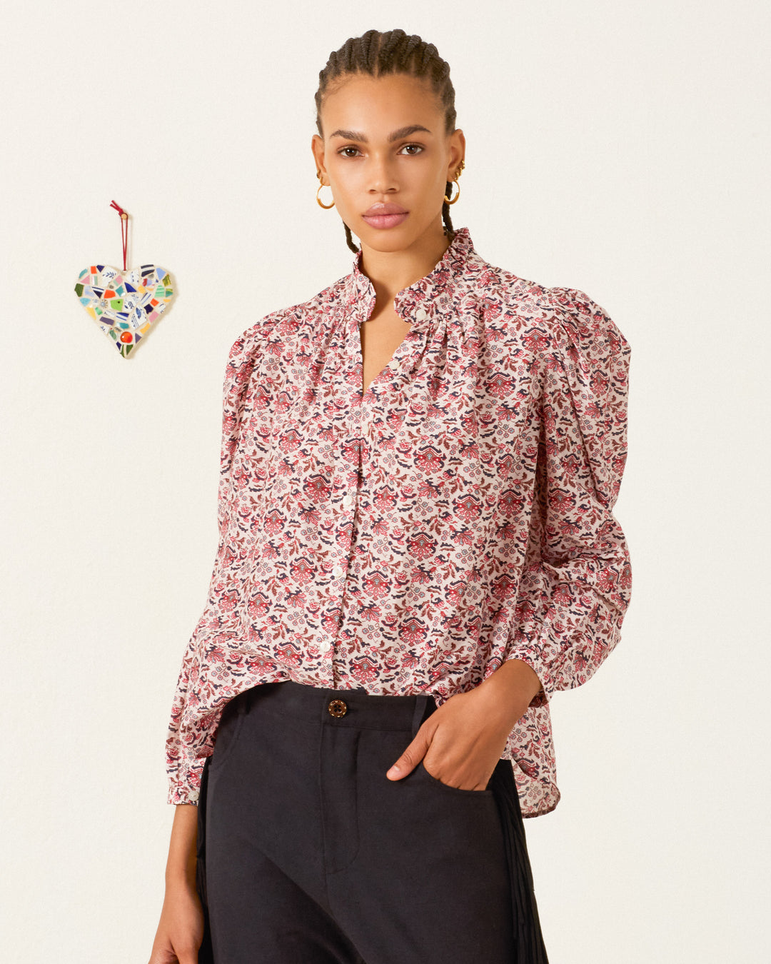 Woman wearing the Annabel Horizon Kilim Shirt and black trousers stands in front of a plain wall with a colorful mosaic heart decoration hanging on it.