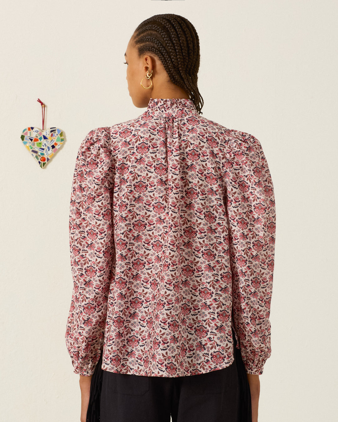 A person stands with their back to the camera, wearing the Annabel Horizon Kilim Shirt—a long-sleeved top featuring a floral kilim pattern, puffed shoulders, and a subtle ruffled collar. To the left on the off-white wall hangs a small multicolored mosaic heart decor.