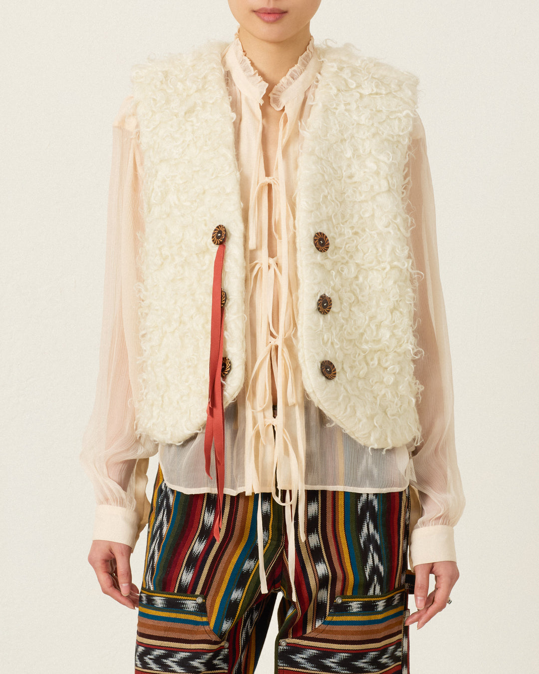 Wearing the Arizona Eventide Curl Vest—a cream-colored, hand-loomed cotton ikat piece—layered over a sheer blouse with ribbon ties and paired with colorful, patterned pants.