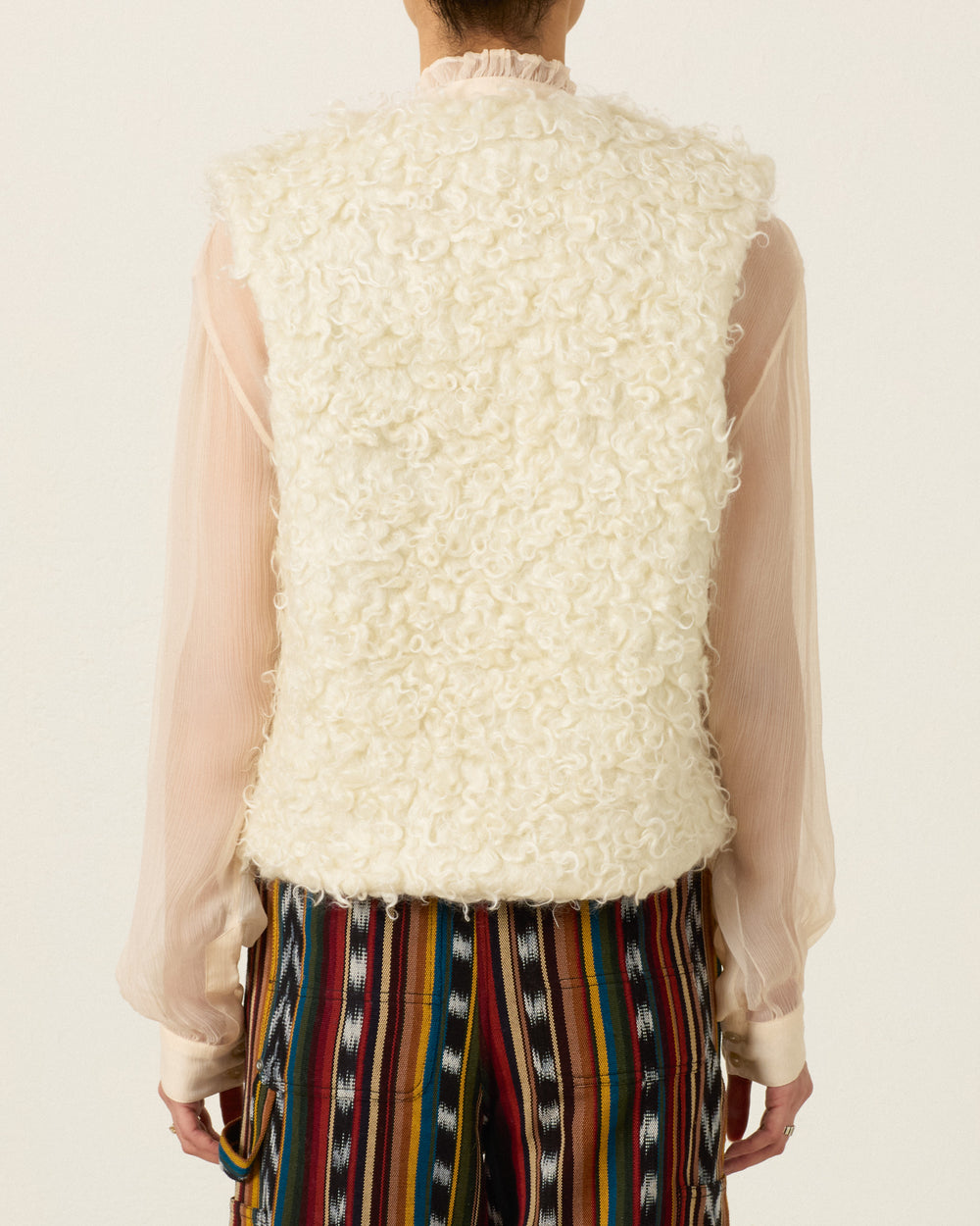 Person wearing the Arizona Eventide Curl Vest— a sleeveless, textured cream Italian mohair piece— over a sheer, long-sleeve blouse with multicolored striped pants, shown from the back.