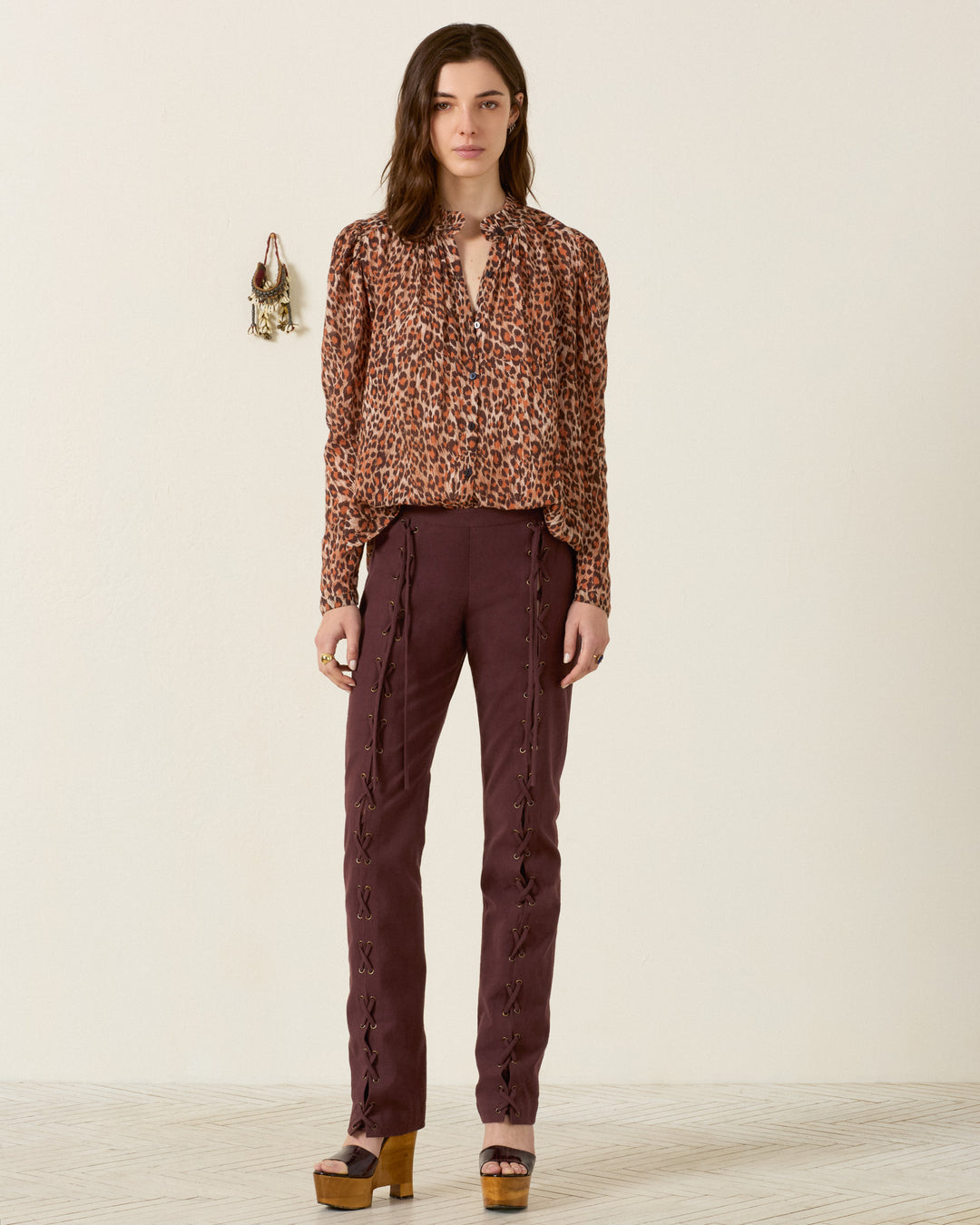 A woman stands against a plain wall wearing the Artemis Leopard Trek Shirt, featuring a ruffled collar. She pairs it with purple pants that have side lacing and platform sandals. A small bag hangs on the wall behind her.