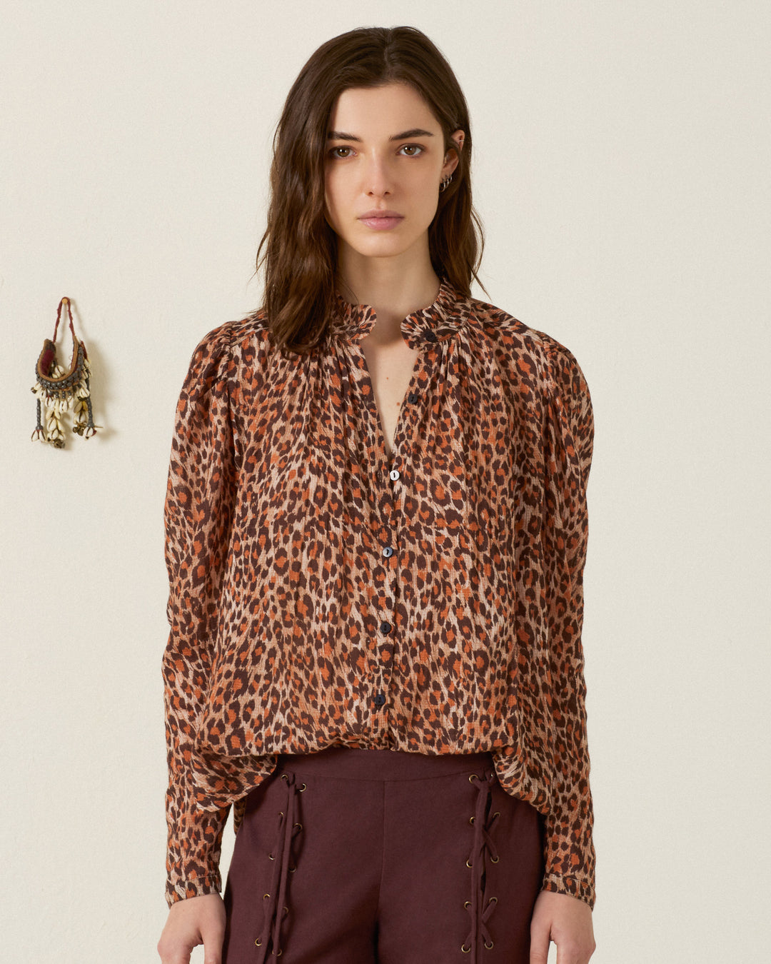 A woman stands against a plain background, wearing the Artemis Leopard Trek Shirt with long puff sleeves, paired with dark brown pants featuring lace-up sides.