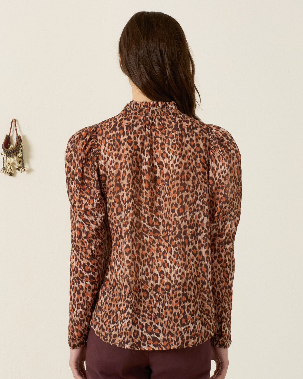 A person stands with their back to the camera, wearing the Artemis Leopard Trek Shirt—a long-sleeve, leopard-print top with puff sleeves—paired with burgundy pants. The cream-colored wall features a small hanging decoration on the left.