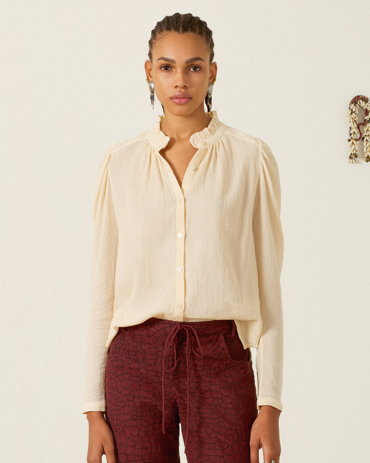 Person standing against a neutral background, wearing the Artemis Prairie Tint Shirt—a light cotton dobby shirt featuring puff sleeves and a ruffled collar—paired with maroon patterned trousers.