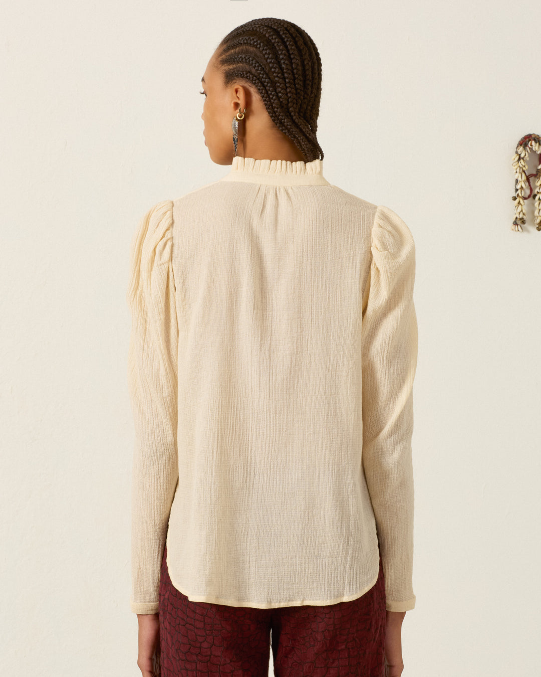 A person with braided hair is seen from the back, wearing the Artemis Prairie Tint Shirt—a long-sleeved, cream-colored cotton dobby top with puff sleeves—paired with dark red pants and standing against a plain light background.