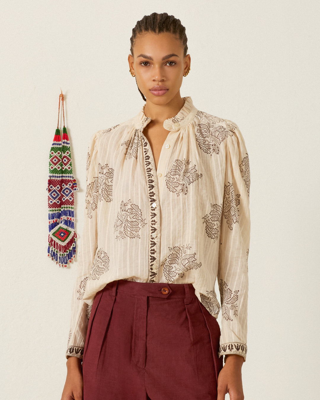 A woman stands against a plain wall, wearing the Artemis Grapevine Stripe Shirt and maroon trousers. A beaded wall hanging is visible to her left.