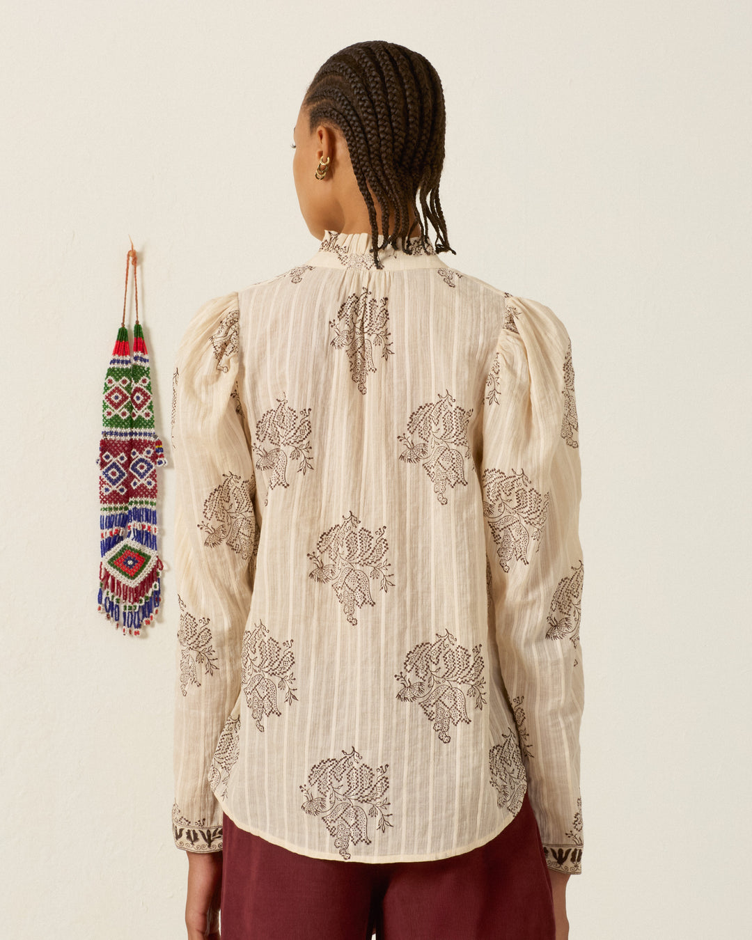 A person with braided hair faces away, wearing the Artemis Grapevine Stripe Shirt—a cream long-sleeve cotton dobby top with dark floral patterns and a subtle ruffled collar—paired with maroon pants. A colorful beaded ornament hangs on the wall to the left.