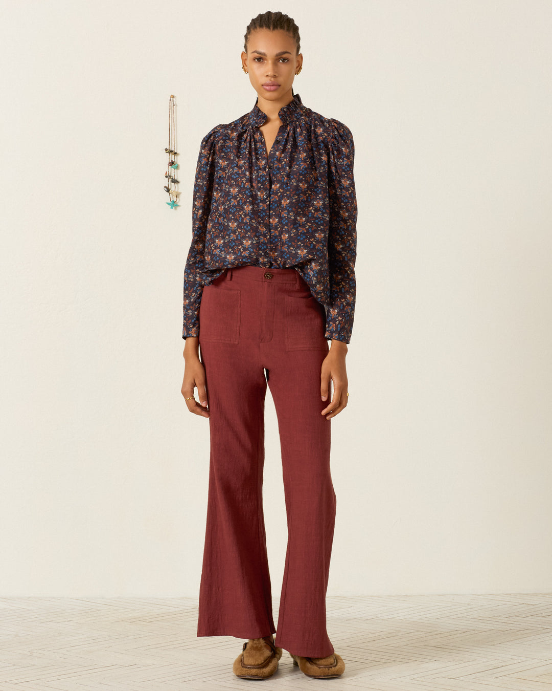 Person stands indoors wearing the Artemis Indigo Kilim Shirt, which features a ruffled collar and puff sleeves in a navy floral pattern, paired with high-waisted maroon pants and brown shoes. Their hair is in braids, and jewelry hangs on the wall behind them.