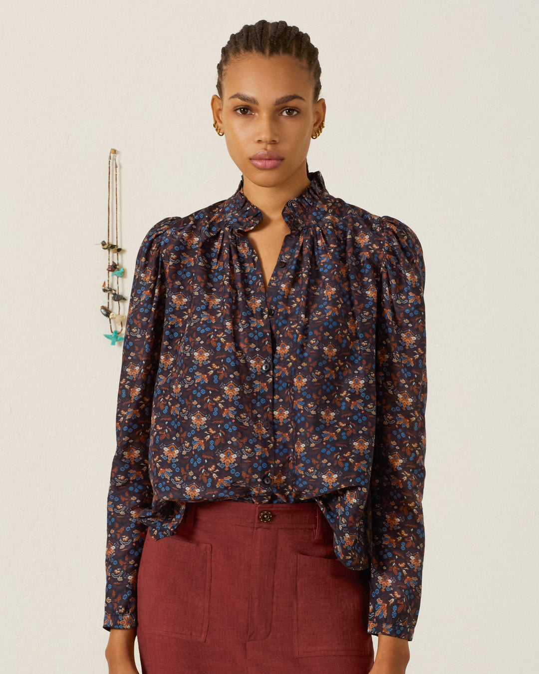 A person stands against a plain wall, wearing the Artemis Indigo Kilim Shirt paired with maroon pants. Several necklaces hang on the wall in the background.