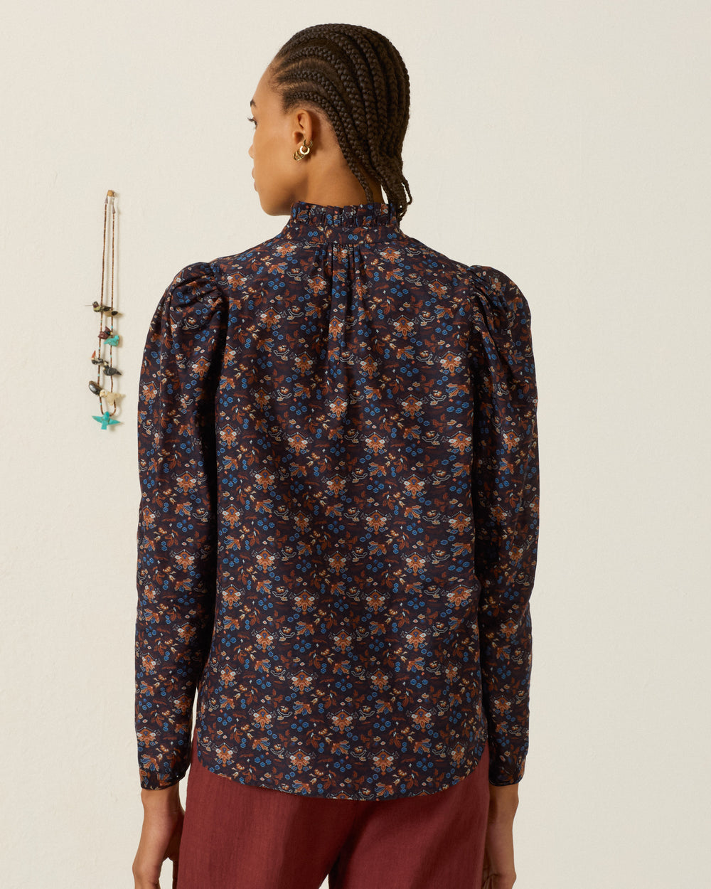 A person with braided hair wears the Artemis Indigo Kilim Shirt—a high-neck, long-sleeve top with a ruffled collar and puff sleeves—paired with red pants, standing with their back to the camera near a wall hanging.