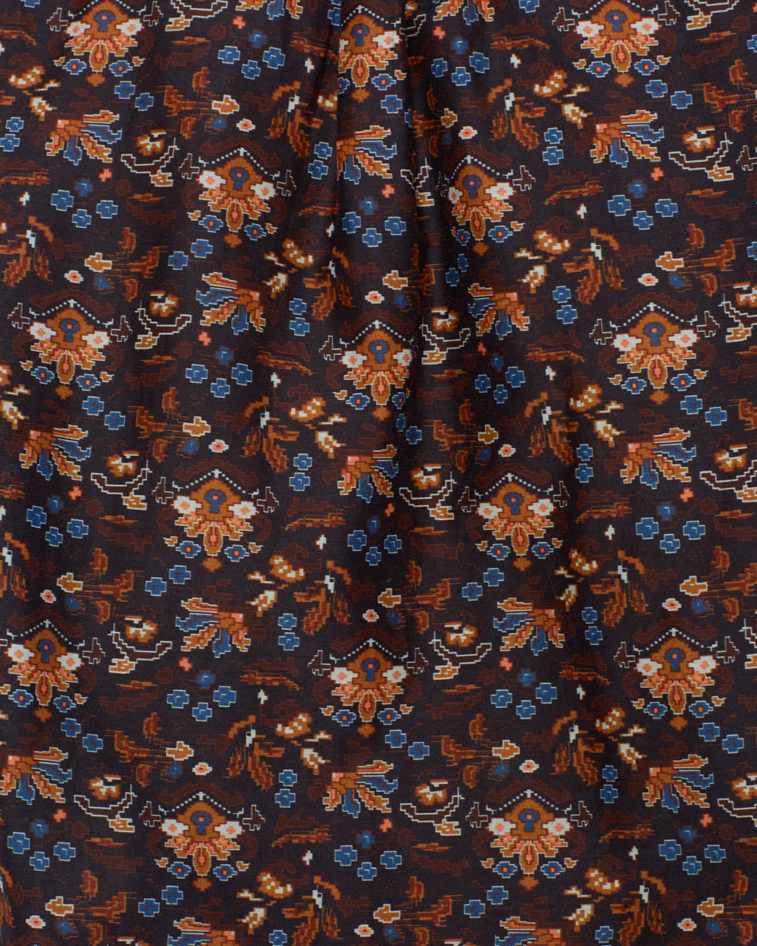Close-up of the Artemis Indigo Kilim Shirt, crafted from a cotton-silk blend fabric featuring a repeating floral and geometric pattern in orange, blue, and beige on a dark background. The fabric appears slightly gathered at the top center, hinting at subtle puff sleeves or a ruffled collar detail.