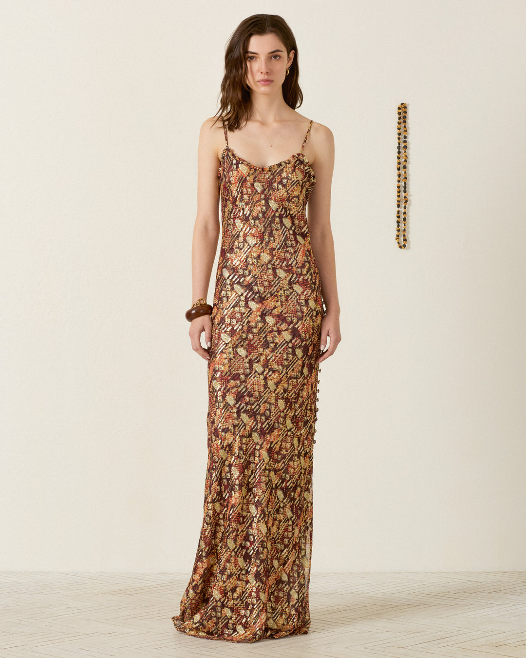 Woman stands facing forward wearing the Athens Topaz Snakeskin Dress, a sleeveless, floor-length slip dress with thin straps; she wears a bracelet on her wrist and a beaded necklace hangs on the wall behind her.