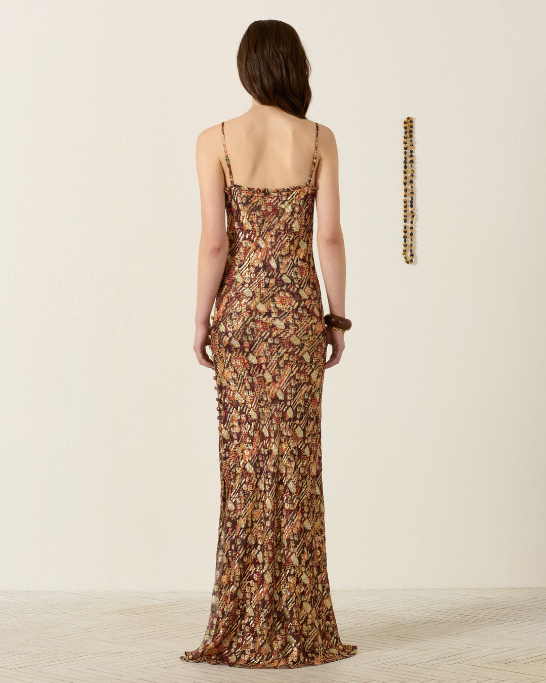 A woman stands facing away, wearing the long, fitted Athens Topaz Snakeskin Dress with thin straps. A beaded necklace hangs on the wall to her right.