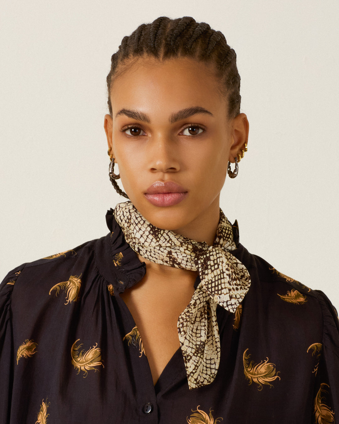 A woman with braided hair, gold hoop earrings, and a patterned black floral shirt faces the camera against a plain background, wearing the Bodhi Lemon Snakeskin Bandana—crafted from silky modal with hand-rolled edges—tied around her neck.
