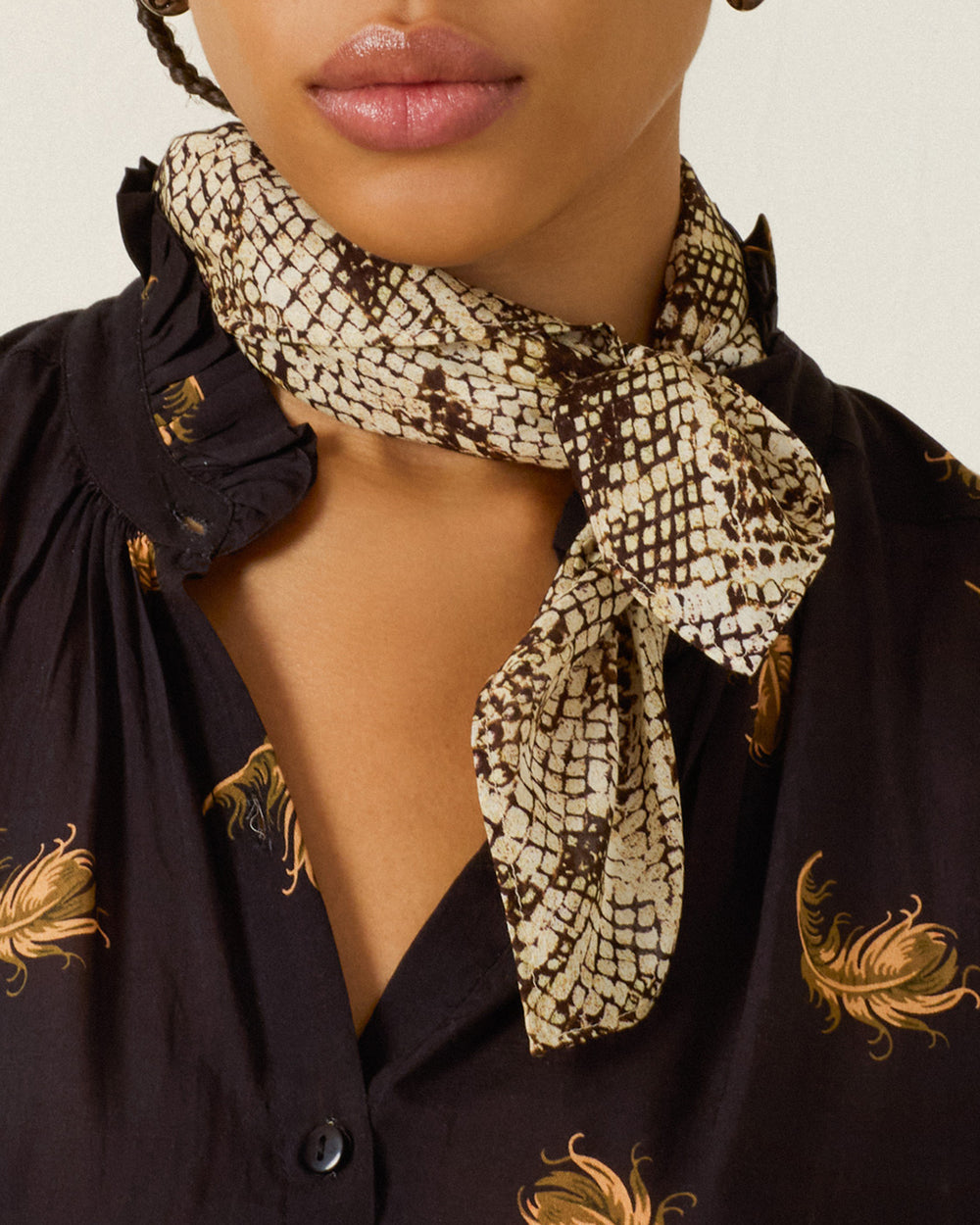 A close-up of a person wearing the Bodhi Lemon Snakeskin Bandana with hand-rolled edges tied around their neck and a black shirt featuring a yellow feather print.