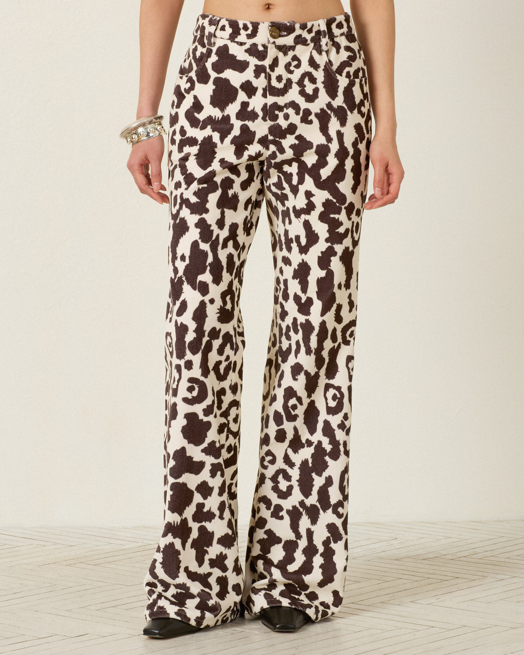 A person wearing the Bunny Leopard Steppe Jean, featuring a high-waisted, flared-leg silhouette in a brown and white leopard print crafted from soft cotton velvet; black shoes are visible.