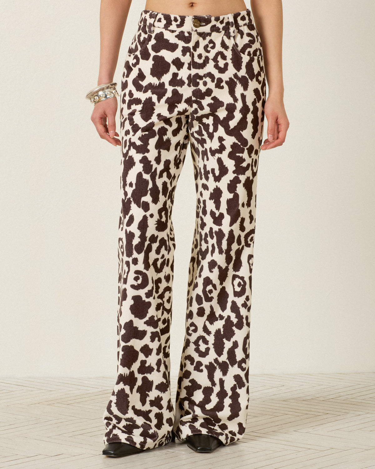 A person wearing the Bunny Leopard Steppe Jean, featuring a high-waisted, flared-leg silhouette in a brown and white leopard print crafted from soft cotton velvet; black shoes are visible.