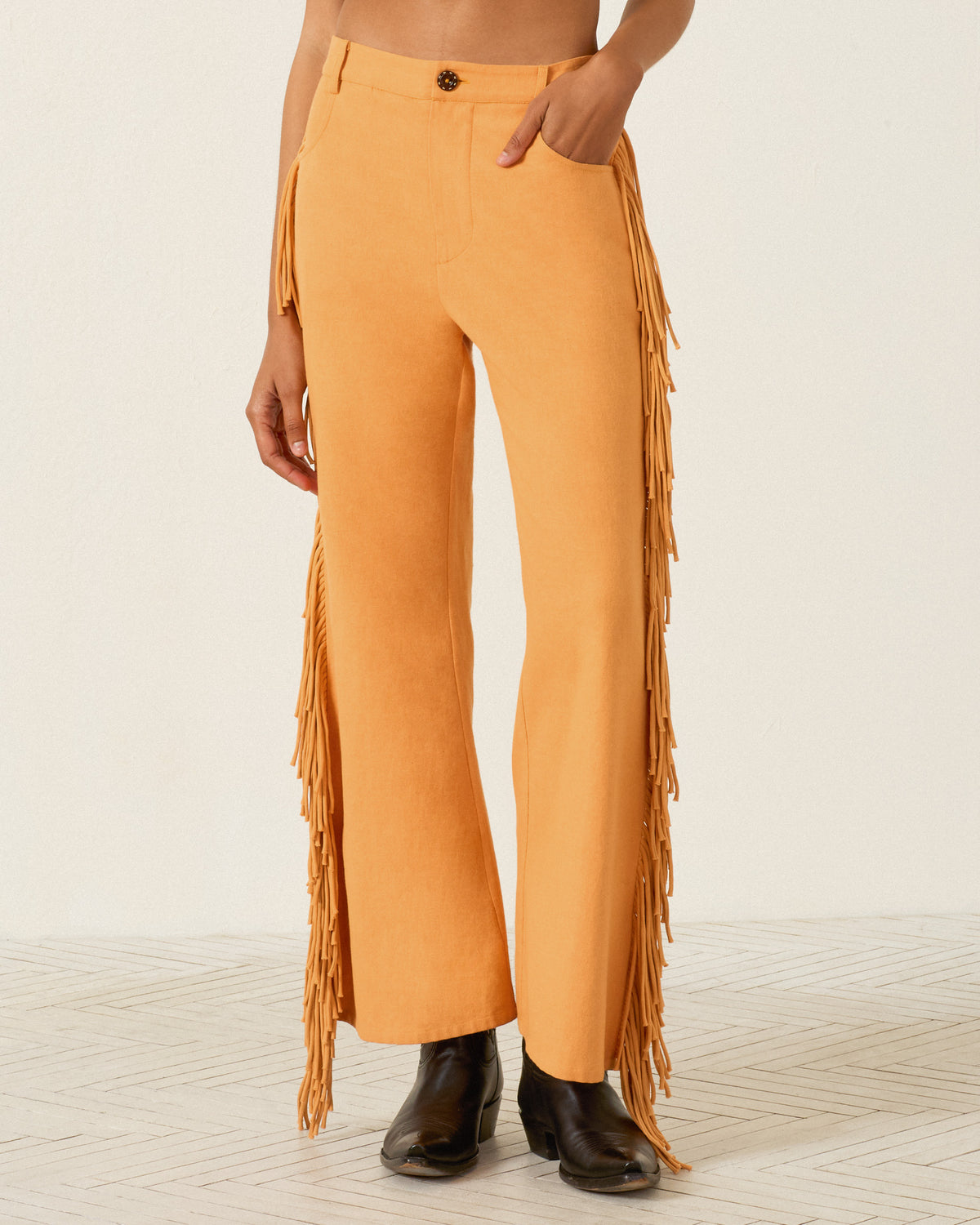 A person wearing the Bunny Sunset Fringe Pant—high-waisted, Western-inspired orange pants with fringe along the sides and a flared leg—paired with black boots and standing on a light patterned floor.