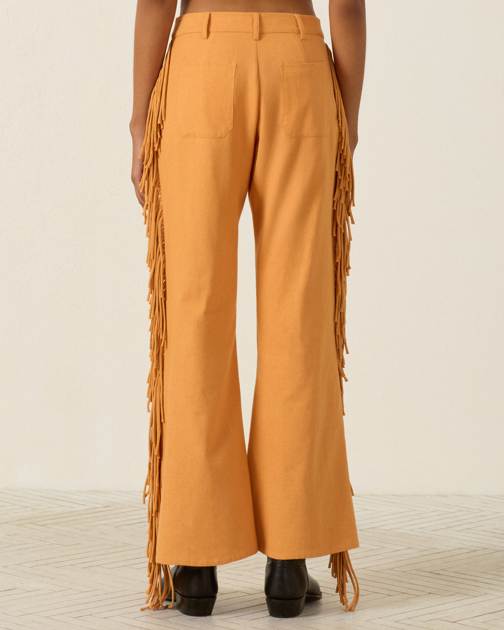 A person is seen from the back standing on a light patterned floor, wearing the Bunny Sunset Fringe Pant—high-waisted, Western-inspired orange pants featuring fringe details along the sides and a flared leg silhouette.