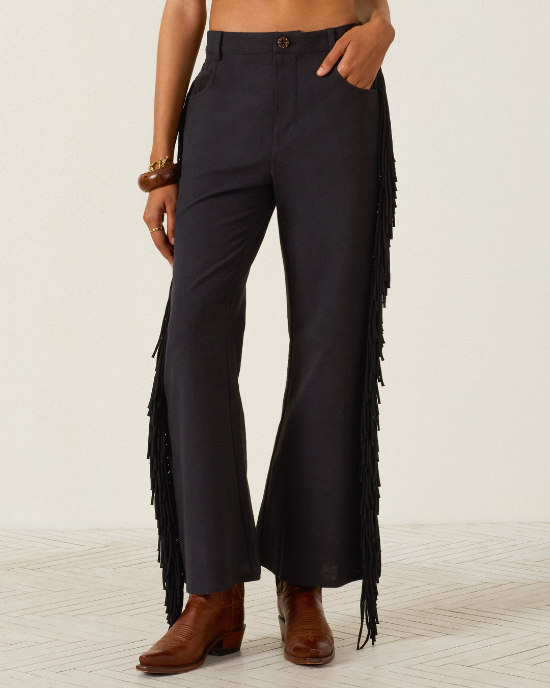 A person wearing the Bunny Onyx Fringe Pant—black, Western-inspired wide-leg pants with fringe on the sides—paired with brown ankle boots and a brown bracelet, standing against a plain background.