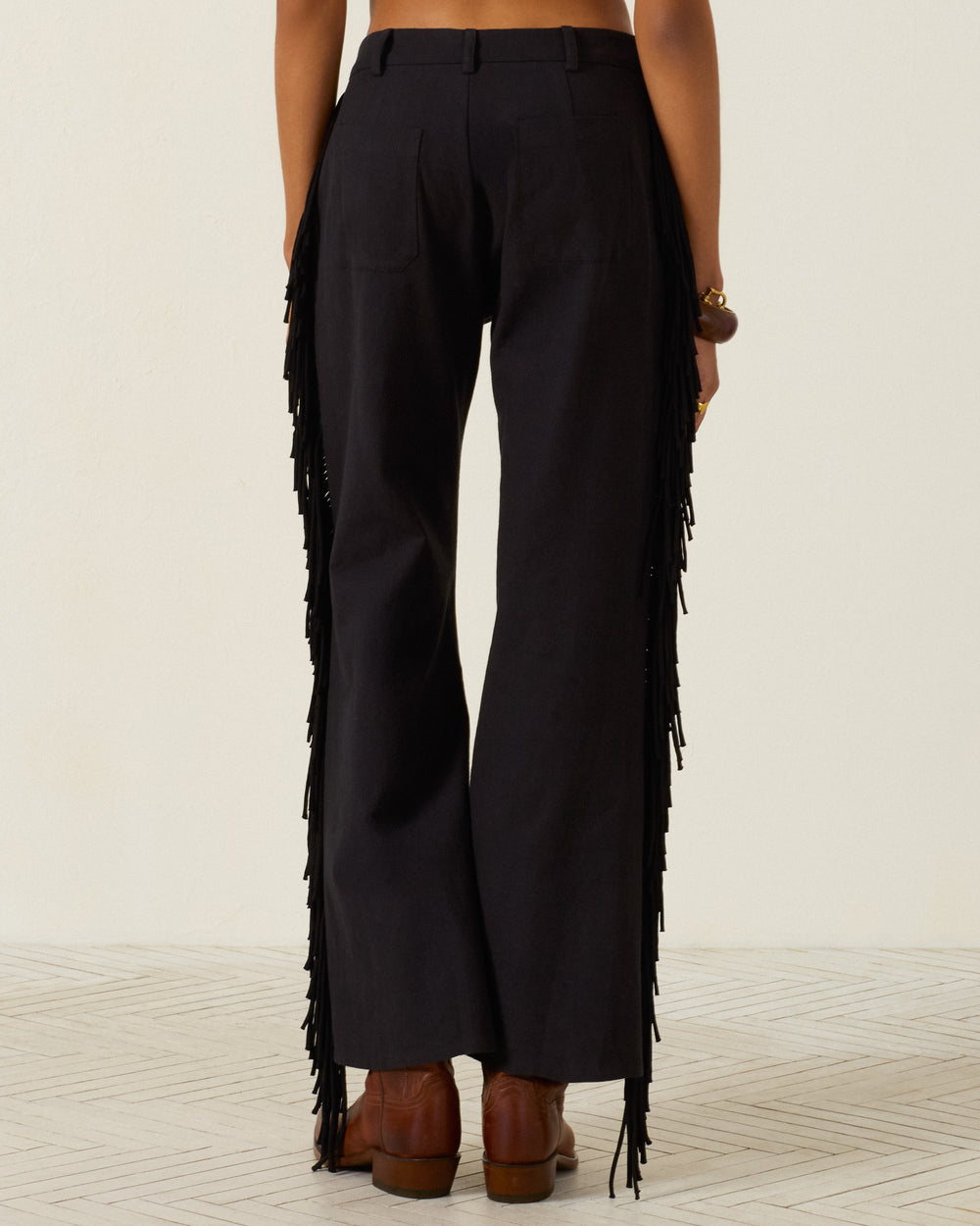 A person is shown from the back, standing on a light patterned floor, wearing the Bunny Onyx Fringe Pant—black, Western-inspired pants featuring fringe along the sides—paired with brown heeled shoes and a bracelet.