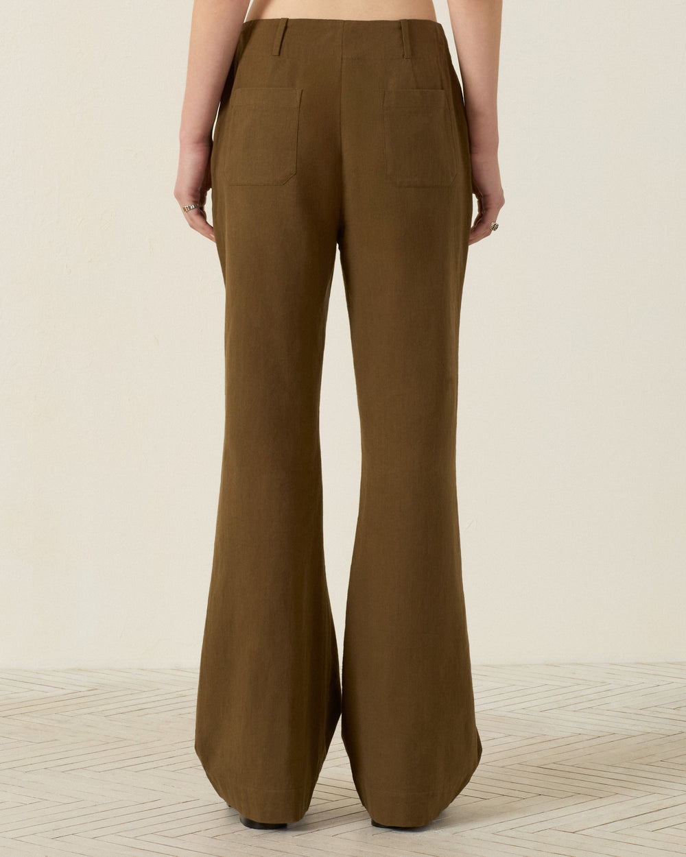 A person is seen from the back wearing the Charlie Olive Branch Pant—high-waisted, wide-leg trousers crafted from khadi cotton in a brown hue—standing on a light-colored floor against a plain wall.