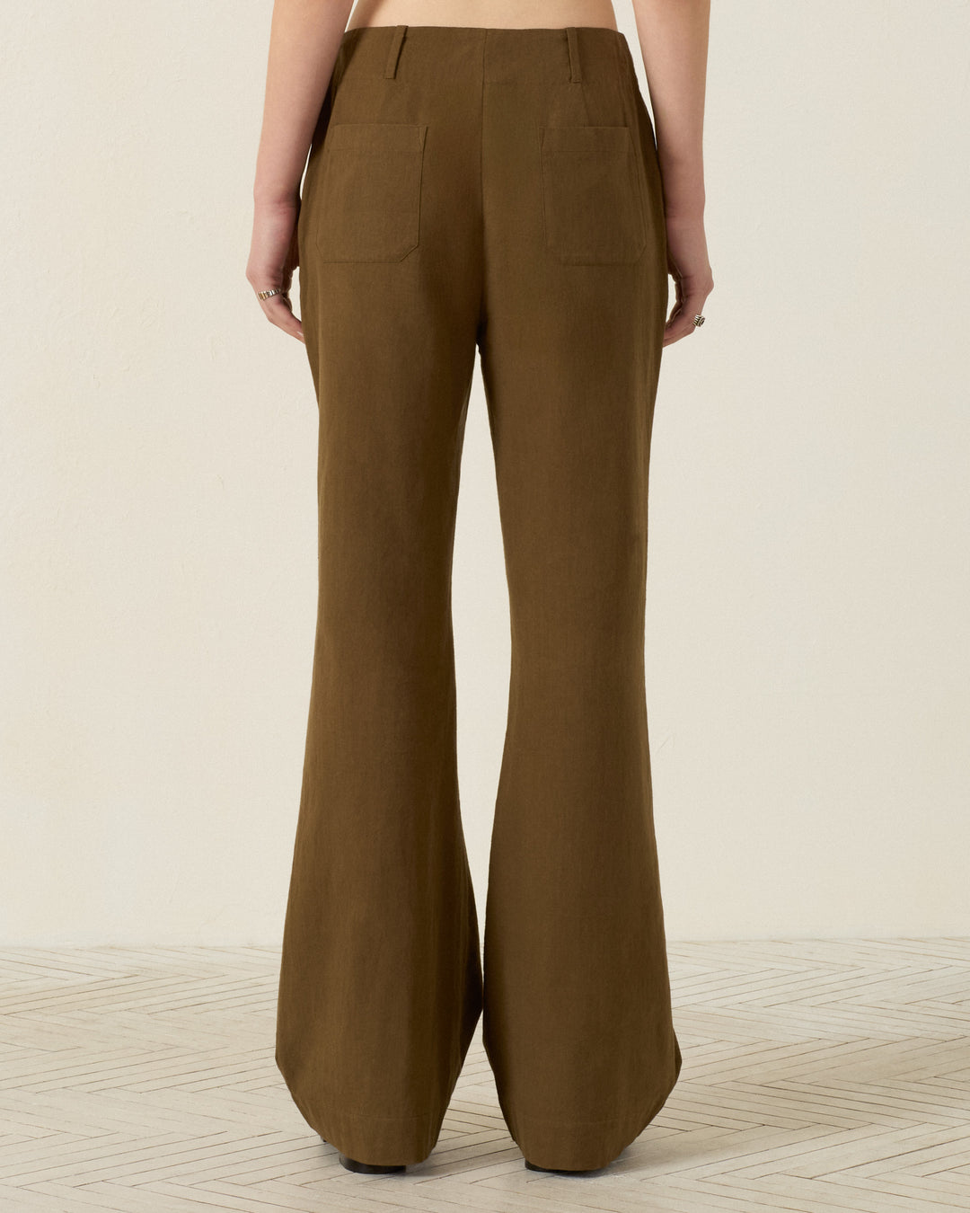 A person is seen from the back wearing the Charlie Olive Branch Pant—high-waisted, wide-leg trousers crafted from khadi cotton in a brown hue—standing on a light-colored floor against a plain wall.