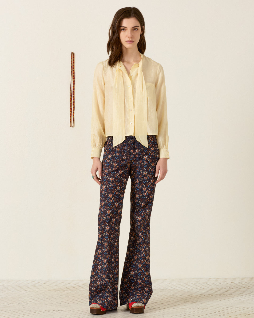A person stands indoors wearing a cream blouse and the Cooper Indigo Kilim Pant—dark floral flared-leg pants—paired with red shoes, while a beaded necklace hangs on the wall behind them.