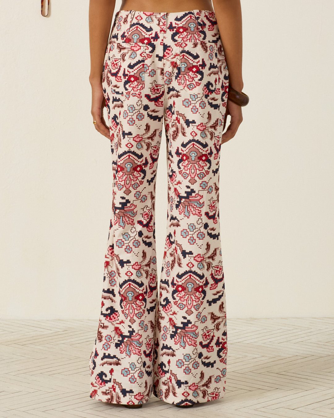 Wearing the Charlie Horizon Kilim Pant—high-waisted flared pants featuring a kilim-inspired pattern in red, black, and cream—a person stands on a light-colored textured floor, viewed from the back.