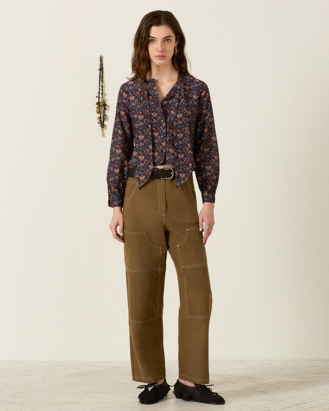 Woman standing indoors wearing the Molly Indigo Kilim Shirt, a navy cotton-silk blouse featuring kilim-inspired motifs, styled with khaki wide-leg pants with visible stitching, a black belt, and black fringed shoes. A necklace hangs on the wall behind her.