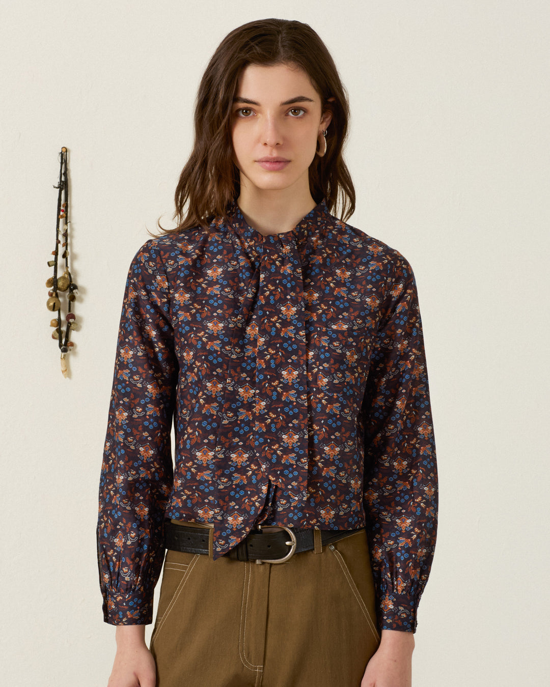 Wearing the Molly Indigo Kilim Shirt—a long-sleeve cotton-silk blouse adorned with kilim-inspired motifs—a woman with wavy brown hair pairs it with khaki pants and a black belt, standing against a plain light wall with a hanging ornament in the background.