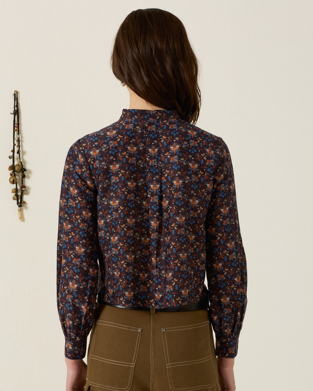 A person stands facing away, wearing the Molly Indigo Kilim Shirt—a long-sleeve, dark printed blouse crafted from a luxurious cotton-silk blend—and brown pants with visible back pockets. On the left side of the image, a decorative wall hanging with kilim-inspired motifs is visible.