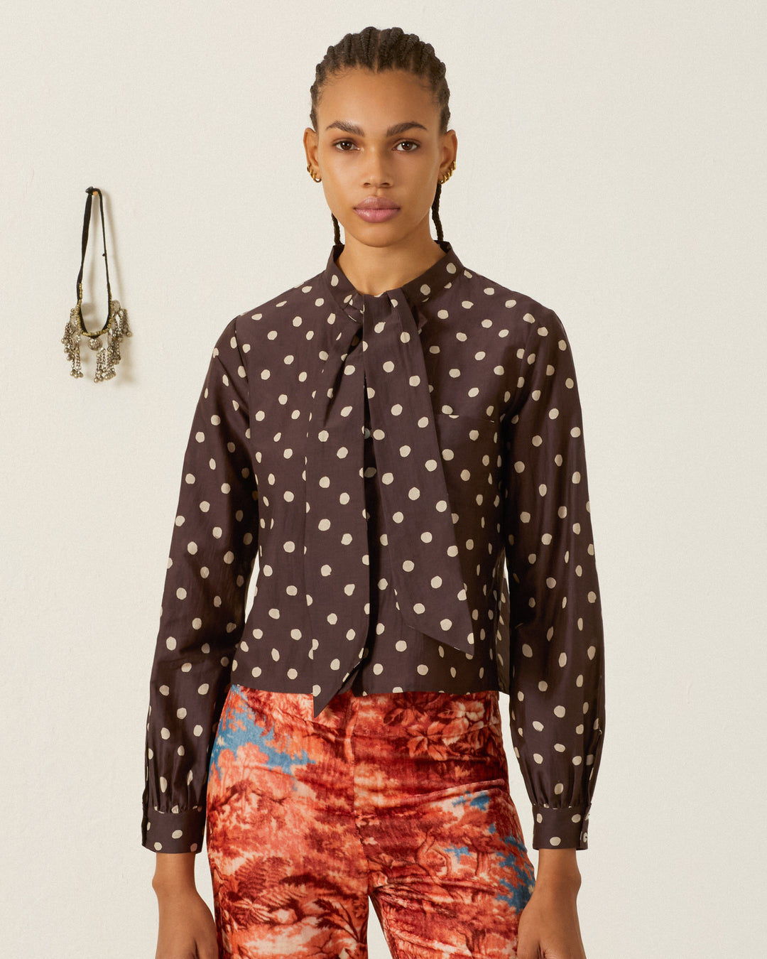 A person wearing the Molly Mahogany Dot Shirt stands in front of a plain wall adorned with hanging jewelry, paired with red patterned pants.