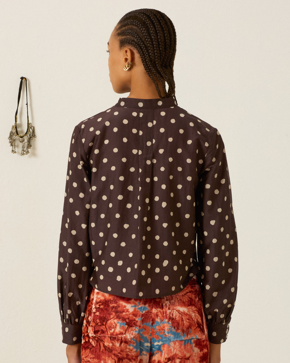 A person is standing indoors with their back to the camera, wearing the Molly Mahogany Dot Shirt—a black blouse with white polka dots made from a cotton-silk blend—and red patterned pants. Wall decor featuring jewelry can be seen on the left side.