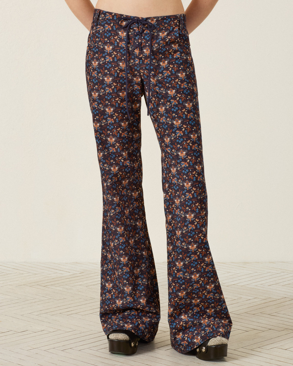 Wearing the Cooper Indigo Kilim Pant, which features a high-waisted fit, flared legs, a small floral print, and a drawstring waistband, a person stands on a light patterned floor with platform shoes.