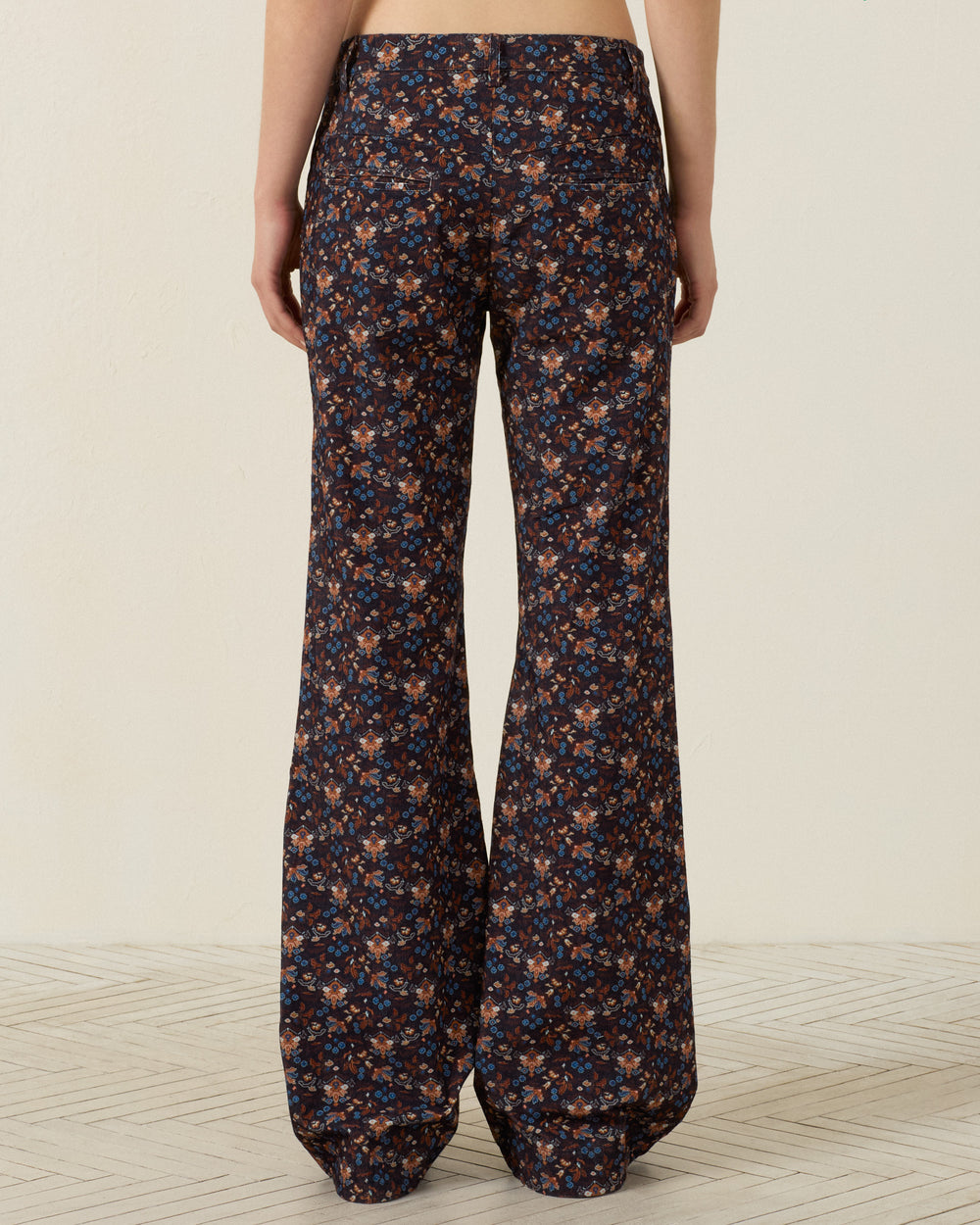 A person is standing with their back to the camera, wearing the Cooper Indigo Kilim Pant—a high-waisted, flared leg style featuring a dark floral pattern. The background shows a plain light-colored wall and a geometric floor.