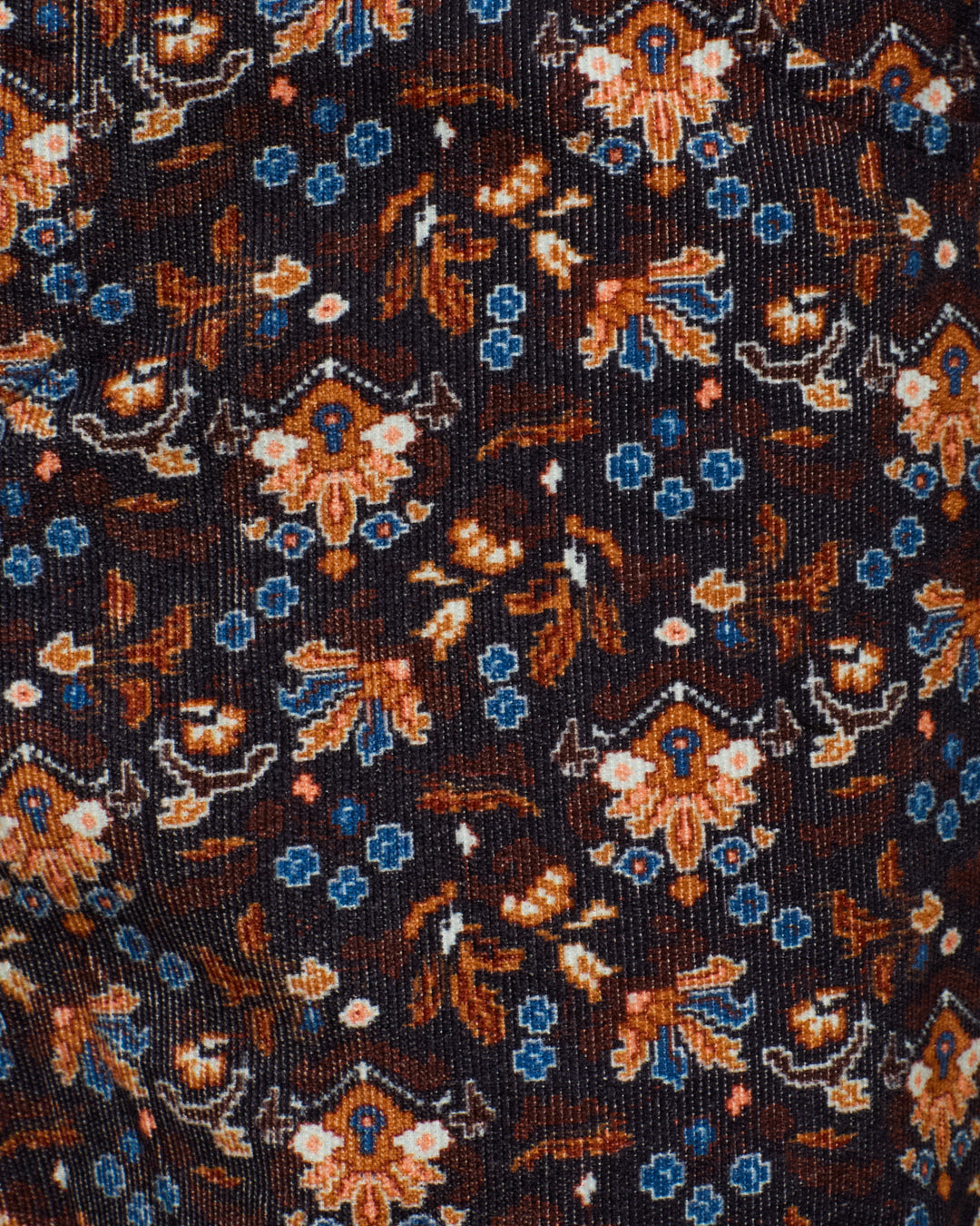 Close-up of the Cooper Indigo Kilim Pant fabric with a dark background, featuring a repeating floral pattern in brown, blue, and cream tones inspired by traditional kilim motifs.