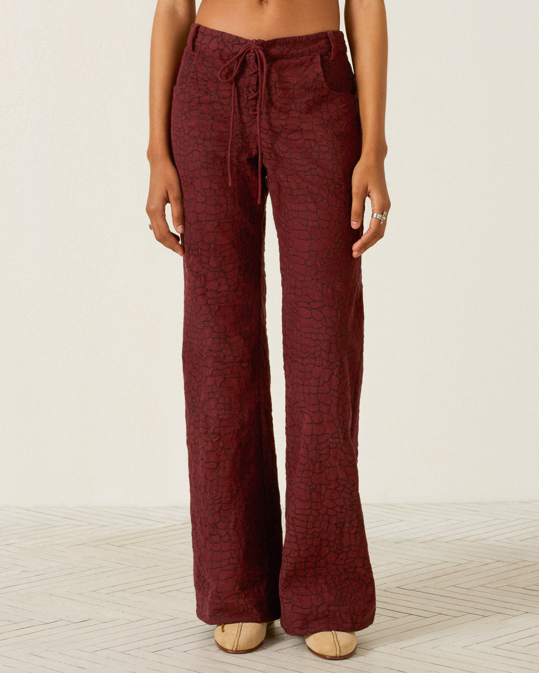 Wearing the Cooper Auburn Crocodile Pant—high-waisted and textured in a rich maroon corduroy with lace-up detailing, drawstring waist, front pockets, and a subtle flared leg—the person stands on a light patterned floor against a plain background.